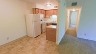 This single-floor residential space offers a practical and comfortable layout designed for everyday living. The kitchen is a standout feature, showcasing warm honey-toned wood cabinetry that provides ample storage, complemented by light-colored countertops and a full suite of white appliances including a refrigerator, electric range, and dishwasher. A built-in sink with under-cabinet lighting adds functionality, while the peninsula-style partial wall creates a natural division between the kitchen and the adjacent living areas. The kitchen is finished with durable, neutral-toned tile flooring that is both easy to maintain and visually clean. Transitioning from the kitchen, a carpeted hallway extends toward the remaining rooms, offering a softer underfoot feel throughout the living spaces. Flush-mount ceiling lighting provides consistent illumination throughout, maintaining a bright and welcoming atmosphere. The overall layout is efficient and well-organized, making the most of the available space. The neutral color palette of soft greens and whites on the walls contributes to an open, airy ambiance, making this a practical and inviting home well-suited for comfortable daily living.