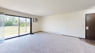 Located in Woodbury, Minnesota, this single-floor 2-bedroom, 2-bathroom residence offers a practical and comfortable layout. The kitchen is well-appointed with white cabinetry, granite-style countertops, wood-look flooring, a stainless steel dishwasher, and white appliances including a refrigerator and electric range — all connected via an open pass-through to the dining area, which features a classic chandelier overhead. The spacious living room is carpeted and filled with natural light from a large sliding glass door that opens to a view of lush greenery, and includes a wall-mounted air conditioning unit and baseboard heating. Both bedrooms are carpeted and generously sized, with the primary bedroom offering its own wall AC unit and a large window overlooking outdoor greenery. The second bedroom features a wide sliding closet and access to the hallway. A carpeted hallway with dark wood-toned doors connects the bedrooms to the two bathrooms, one of which includes a full tub-shower combination with tile surround and stone-look tile flooring. A dedicated closet provides additional storage. The overall ambiance is clean, neutral, and bright throughout.