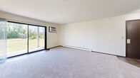 Located in Woodbury, Minnesota, this single-floor 2-bedroom, 2-bathroom residence offers a practical and comfortable layout. The kitchen is well-appointed with white cabinetry, granite-style countertops, wood-look flooring, a stainless steel dishwasher, and white appliances including a refrigerator and electric range — all connected via an open pass-through to the dining area, which features a classic chandelier overhead. The spacious living room is carpeted and filled with natural light from a large sliding glass door that opens to a view of lush greenery, and includes a wall-mounted air conditioning unit and baseboard heating. Both bedrooms are carpeted and generously sized, with the primary bedroom offering its own wall AC unit and a large window overlooking outdoor greenery. The second bedroom features a wide sliding closet and access to the hallway. A carpeted hallway with dark wood-toned doors connects the bedrooms to the two bathrooms, one of which includes a full tub-shower combination with tile surround and stone-look tile flooring. A dedicated closet provides additional storage. The overall ambiance is clean, neutral, and bright throughout.