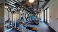 This fitness center offers a comprehensive workout environment designed to accommodate a variety of exercise routines. The space is organized into distinct functional zones, beginning with a dedicated cardio area lined with multiple treadmills, elliptical machines, and cable machines, all equipped with individual media screens. Full-length mirrors run along the walls, visually expanding the space and allowing users to monitor their form during exercise. The industrial-style exposed ceiling features dark-painted ductwork and structural elements, complemented by pendant lighting that provides a focused, functional ambiance throughout. Large windows along the perimeter allow natural light to fill the space, with views of an outdoor greenery area adding a refreshing visual element. The strength-training section is well-appointed with a multi-tiered dumbbell rack offering a full range of weights, adjustable benches, and cable machines suitable for a wide range of exercises. A glass-enclosed room is accessible from the main floor, offering a more private workout area. The flooring transitions between durable hardwood-style material in the cardio zone and cushioned fitness flooring in the strength area, providing appropriate support for each type of training.