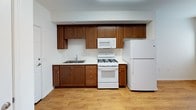 This single-floor unit offers a clean, functional layout with a cohesive neutral color scheme and warm hardwood-style flooring that runs throughout. The kitchen is a practical highlight, featuring rich medium-toned wood cabinetry extending to the upper walls for generous storage capacity. White appliances — including a gas range, over-the-range microwave, and full-size refrigerator — complement the light countertops and stainless steel sink. The open sightlines connecting the kitchen to adjacent areas create an easy flow between spaces.

A louvered closet door and a paneled entry door are positioned conveniently off the kitchen area, offering accessible storage and a direct entryway. The bathroom includes a full tub and shower combination with white tile surround, chrome fixtures, and a built-in wall shelving unit for added convenience.

The bedroom is bright and simply designed, with a large window fitted with horizontal blinds that allow for natural light while maintaining privacy. Crisp white walls and trim throughout the unit contribute to an airy, well-maintained atmosphere. The overall design prioritizes functionality and ease of living within a tidy, well-organized space.