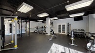 This fitness center is a thoughtfully designed, multi-zone facility built to support a wide range of workout styles. The space is organized into distinct areas, each tailored to a specific type of training. The cardio zone features a row of treadmills and stationary bikes, positioned near large floor-to-ceiling windows that flood the area with natural light and offer views of the surrounding outdoors. The strength training area is outfitted with a power rack, adjustable benches, dumbbells, medicine balls, and a cable machine, providing comprehensive free weight and resistance training options. A dedicated functional training zone offers an open, flexible layout with plyo boxes, push sleds, and ample open floor space for dynamic movement and group workouts. Frosted glass partition panels divide sections of the space, providing visual separation while maintaining an open, airy feel. Throughout the facility, dark rubber flooring provides durability and traction, while an exposed black ceiling with industrial-style suspended lighting fixtures gives the space a modern, athletic aesthetic. Structural columns punctuate the layout, and a wall-mounted television is positioned in the functional training area. French doors connect zones within the facility, enhancing flow and accessibility across the entire center.
