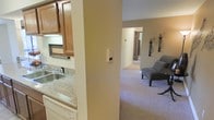 This single-floor residence features a practical, open layout that connects the kitchen and living areas seamlessly. The kitchen is equipped with warm oak-toned cabinetry, a granite-look countertop, a stainless steel sink, and a dishwasher, offering functional workspace for everyday use. A structural column partially divides the kitchen from the adjoining living space, maintaining a sense of openness while subtly defining each area.

The living area is finished with wall-to-wall neutral carpeting that contributes to a warm, comfortable atmosphere. A modern lounge chair provides seating, and wall-mounted decorative accents add a touch of personality to the space. A hallway visible in the background suggests access to additional rooms, indicating a multi-room floor plan with private spaces beyond the main living zone.

Throughout the residence, the walls are painted in soft, neutral beige tones that complement the carpeting and cabinetry, creating a cohesive and understated aesthetic. The layout prioritizes ease of movement between the kitchen and living areas, making the space well-suited for everyday living.