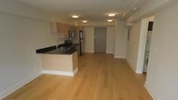 This single-floor space features a clean, modern design centered around an open-concept layout that seamlessly connects the kitchen and living areas. The light hardwood flooring extends throughout, lending warmth and continuity to the space. The kitchen is well-appointed with light wood cabinetry, dark granite countertops, and a full suite of stainless steel appliances including a refrigerator, range, and microwave. A kitchen island with a built-in sink serves as both a functional workspace and a natural divider between the cooking and living zones. Flush-mount ceiling lighting provides consistent, even illumination throughout. The open living area offers a generous, flexible space with clean white walls and a minimalist aesthetic. A solid entry door and a structured layout contribute to the unit's sense of privacy and practicality. The overall ambiance is contemporary and efficient, with thoughtful design choices that maximize the use of the available space while maintaining a bright, welcoming atmosphere.