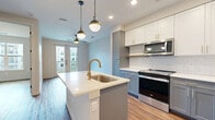 This thoughtfully designed one-bedroom, one-bathroom residence in Austin, TX offers a cohesive open-concept layout that maximizes every square foot. The kitchen serves as the centerpiece of the living area, showcasing a large center island with a white quartz countertop and a gold-tone undermount sink. Two stylish pendant lights hang above the island, complementing the two-tone cabinetry — white upper cabinets with gold hardware paired with slate-blue lower cabinets. A classic white subway tile backsplash, stainless steel appliances including a gas range, over-the-range microwave, dishwasher, and a full-size side-by-side refrigerator complete the kitchen. Warm hardwood-style flooring runs throughout the entire unit, creating a unified and inviting feel. The bedroom is a bright, comfortable retreat with a large window that brings in natural light and offers views of the surrounding neighborhood. A dedicated closet provides practical storage. The bathroom is elegantly finished with a gray vanity topped with white quartz, gold-accented fixtures, a large mirror, and a glass-enclosed walk-in shower with floor-to-ceiling tile. The unit's clean, neutral color palette and modern gold and black hardware accents give it a polished, contemporary character.