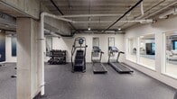 This fitness center offers a comprehensive workout environment spread across a single level, designed to accommodate a wide range of exercise needs. The space features an industrial aesthetic, with exposed concrete ceilings, visible ductwork and piping, and track lighting that lends the facility a modern, urban character.

The cardio zone is equipped with multiple treadmills and a stair climber machine, all positioned along mirrored walls that enhance the sense of openness. A dedicated strength training area houses a multi-station cable and weight machine alongside a barbell setup, providing versatile options for resistance training. A rack of dumbbells in varying weights rounds out the free weight offerings.

A separate functional training area contains stability balls in multiple sizes, medicine balls, resistance bands, exercise mats, and a compact set of orange lockers for personal storage. An elliptical trainer is also present in this section, adding further cardio variety.

Large interior windows along one wall look into an adjacent lounge or common area furnished with seating arrangements and exposed brick accents, creating a visual connection between the fitness space and the broader amenity floor. The rubber-speckled flooring throughout provides cushioning and durability, while recessed and track lighting ensures the entire space is well-lit and functional.