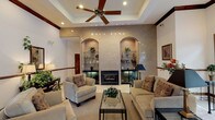 This single-floor clubhouse offers a refined and welcoming atmosphere designed for community gathering and relaxation. The grand entrance foyer sets the tone with polished marble tile flooring, rich dark wood trim, decorative columns, and expansive French doors framed by transom windows that flood the space with natural light and offer views of the outdoor pool area. A pair of cream leather armchairs and wall-mounted sconces add a classic, inviting touch to this transitional space.

The main lounge is a standout feature of the clubhouse, centered around a built-in fireplace flanked by two elegant arched display niches with glass shelving, all set against a textured, shimmering accent wall. A coffered tray ceiling with a large ceiling fan and recessed lighting crowns the room, while plush neutral sofas and a glass-top coffee table create a comfortable and stylish seating arrangement.

Adjacent to the lounge, a dining and gathering area features a tray ceiling, ceiling fan, and a round glass dining table surrounded by wrought iron chairs. A kitchenette with a dark countertop, bar-height seating, and track lighting provides functional amenities for events and social occasions. Throughout, consistent use of crown molding, warm wood accents, and neutral tones give the clubhouse a cohesive and sophisticated character.