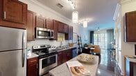 This single-story, one-bedroom, one-bathroom unit offers a thoughtfully designed layout that maximizes its compact footprint. The open-concept kitchen and living area serves as the heart of the space, featuring rich dark hardwood flooring, crown molding, and elevated ceilings that enhance the sense of volume. The kitchen is outfitted with deep-toned wood cabinetry, granite countertops, a central island with pendant lighting, and a full suite of stainless steel appliances, including a refrigerator, electric range, microwave, and dishwasher. Track lighting supplements the natural light flowing in from the living area.

The living room opens directly onto a private balcony through full-height sliding glass doors dressed with floor-length draping curtains, bringing in generous natural light and outdoor views. The bedroom is a cozy retreat featuring carpeted flooring, neutral grey walls, a large window with blinds and curtain panels, and a sliding barn-style closet door for efficient space use.

The bathroom is finished with tile flooring, a dark wood vanity with granite countertop, a large mirror, and a full tub-shower combination enclosed with large-format wall tile. A connecting hallway provides a functional transition between the private and shared areas of the unit.