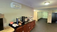 This single-floor space is thoughtfully designed to serve guests with ease and comfort. The focal point of the area is a well-appointed refreshment station, featuring rich wood cabinetry with a dark granite countertop. The station is fully equipped with coffee and tea brewing equipment, thermal dispensers, a variety of beverage condiments, and an assortment of packaged snacks and drink options — catering to a range of preferences. An overhead wire rack provides additional storage and organization for cups and supplies. The space continues into a hallway corridor, illuminated by ceiling-mounted lighting and finished with dark carpeted flooring throughout. The walls are painted in a neutral sage green tone, contributing to a calm and welcoming ambiance. The textured ceiling and practical layout reflect a functional yet inviting design suited for a hospitality or lodging environment. The corridor leads to additional areas of the property, suggesting a broader layout designed to accommodate guests efficiently. Overall, the space balances utility and comfort, making it an accessible and well-organized amenity area.