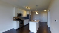 Located at 900 Luxor Lane in Norristown, PA, this single-floor residence showcases a thoughtfully designed open-concept layout that seamlessly blends the kitchen and living areas. The kitchen is a standout feature, appointed with sleek white cabinetry, granite countertops, and a full suite of stainless steel appliances, including a range, microwave, and refrigerator. A central island with an integrated sink and elegant pendant lighting serves as both a functional workspace and a natural gathering point. Dark hardwood flooring runs throughout, creating a rich contrast against the white cabinetry and light-toned walls. Track lighting in the kitchen adds targeted illumination, complementing the warm ambient glow of the pendant fixtures. The open flow into the living area provides a sense of spaciousness and versatility, making the space well-suited for both everyday living and entertaining. Clean lines, contemporary finishes, and a neutral color palette give the residence a modern, polished aesthetic that is both inviting and functional.