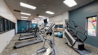 Located at 55 S Vail Ave in Arlington Heights, Illinois, this fitness center offers a thoughtfully equipped exercise space designed to support a wide range of workout routines. The facility is contained on a single floor and features a spacious, open layout with a drop-panel ceiling fitted with recessed fluorescent lighting that keeps the space bright and welcoming throughout.

The gym is outfitted with a comprehensive selection of cardio machines, including Matrix and Spirit brand treadmills, elliptical trainers, a stair climber, and a recumbent stationary bike. For strength training, the space includes a multi-station cable machine, a flat weight bench, free weights, and an assortment of stability and medicine balls. Wall-mounted flat-screen televisions are positioned throughout for entertainment during workouts.

A full-length mirror runs along one wall, accompanied by a wooden ballet barre—adding a functional touch for stretching and balance exercises. The interior is accented by bold teal-painted walls contrasted against neutral white, giving the space an energetic yet clean aesthetic. Textured carpet tiles cover the floor, providing comfort and traction during exercise. A row of interior windows along one wall allows views into an adjacent area, while decorative accent windows add a unique visual element. Ceiling fans provide additional air circulation, completing a practical and inviting fitness environment.