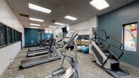 Located at 55 S Vail Ave in Arlington Heights, Illinois, this fitness center offers a thoughtfully equipped exercise space designed to support a wide range of workout routines. The facility is contained on a single floor and features a spacious, open layout with a drop-panel ceiling fitted with recessed fluorescent lighting that keeps the space bright and welcoming throughout.

The gym is outfitted with a comprehensive selection of cardio machines, including Matrix and Spirit brand treadmills, elliptical trainers, a stair climber, and a recumbent stationary bike. For strength training, the space includes a multi-station cable machine, a flat weight bench, free weights, and an assortment of stability and medicine balls. Wall-mounted flat-screen televisions are positioned throughout for entertainment during workouts.

A full-length mirror runs along one wall, accompanied by a wooden ballet barre—adding a functional touch for stretching and balance exercises. The interior is accented by bold teal-painted walls contrasted against neutral white, giving the space an energetic yet clean aesthetic. Textured carpet tiles cover the floor, providing comfort and traction during exercise. A row of interior windows along one wall allows views into an adjacent area, while decorative accent windows add a unique visual element. Ceiling fans provide additional air circulation, completing a practical and inviting fitness environment.