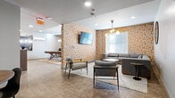 This thoughtfully designed lounge offers a versatile, single-floor layout that seamlessly blends social and recreational spaces. The interior is anchored by a striking warm-toned geometric wallpaper pattern that flows throughout, lending the space a cohesive and contemporary identity.

The main seating area features a generously sized sectional sofa paired with mid-century-inspired accent chairs, all arranged around a round coffee table atop a textured area rug. A wall-mounted flat-screen TV and a modern multi-globe pendant light fixture enhance both function and ambiance in this relaxed gathering zone. Natural light filters in through frosted windows, complementing the recessed ceiling lighting throughout.

Adjacent to the seating area is a dedicated game room centered around a substantial wooden pool table with a classic X-base frame, positioned beneath a matching globe pendant light. A wall-mounted TV and bar-height table with stools provide additional entertainment options in this zone.

The lounge also includes a bar or kitchenette area, visible from the main space, offering counter seating with bar stools for casual dining or socializing. A separate, more intimate meeting room provides a quieter setting with a round wooden table and seating for four, equipped with its own climate control unit.

Durable, neutral-toned flooring runs throughout, tying all areas together in a clean, modern aesthetic suited for community gatherings, recreation, and collaboration.