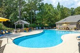 The Cahaba Grandview pool amenity area in Birmingham, Alabama offers a well-appointed outdoor retreat designed for relaxation and recreation. At its centerpiece is a generously sized, freeform swimming pool with clear blue water, accessible via built-in entry steps and handrails. The expansive concrete pool deck surrounds the water on all sides, accommodating multiple lounge chairs and yellow market umbrellas that provide convenient shade throughout the day.

A charming wood-framed gazebo anchors one end of the pool deck, offering a shaded gathering spot. Adjacent to the pool area, a dedicated patio features a handsome stacked-stone fire pit flanked by built-in seating walls, creating an inviting space for evening gatherings. Colorful potted flowers and carefully maintained shrubs add seasonal interest throughout the grounds.

The facility building itself is constructed of classic brick with large multi-pane windows and a covered entrance supported by painted columns with decorative lantern lighting. Manicured landscaping lines the building's perimeter. The entire area is enveloped by mature trees, providing natural privacy and a serene, wooded ambiance that enhances the overall outdoor experience.
