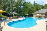 The Cahaba Grandview pool amenity area in Birmingham, Alabama offers a well-appointed outdoor retreat designed for relaxation and recreation. At its centerpiece is a generously sized, freeform swimming pool with clear blue water, accessible via built-in entry steps and handrails. The expansive concrete pool deck surrounds the water on all sides, accommodating multiple lounge chairs and yellow market umbrellas that provide convenient shade throughout the day.

A charming wood-framed gazebo anchors one end of the pool deck, offering a shaded gathering spot. Adjacent to the pool area, a dedicated patio features a handsome stacked-stone fire pit flanked by built-in seating walls, creating an inviting space for evening gatherings. Colorful potted flowers and carefully maintained shrubs add seasonal interest throughout the grounds.

The facility building itself is constructed of classic brick with large multi-pane windows and a covered entrance supported by painted columns with decorative lantern lighting. Manicured landscaping lines the building's perimeter. The entire area is enveloped by mature trees, providing natural privacy and a serene, wooded ambiance that enhances the overall outdoor experience.