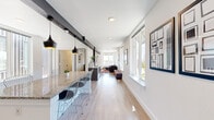 This single-floor space is thoughtfully designed with a contemporary aesthetic, offering an open, elongated layout that maximizes its footprint. Light hardwood flooring runs continuously throughout, lending warmth and visual cohesion to the interior.

At one end, a prominent granite-topped island serves as a central gathering point, surrounded by wire-frame bar stools and illuminated by striking black pendant lights suspended from an exposed structural beam above. Adjacent to the island, a dedicated work area features wall-mounted monitors and seating with vibrant yellow chairs, making it a functional workspace.

The lounge area at the opposite end is bright and welcoming, benefiting from floor-to-ceiling windows that flood the space with natural light and provide open street views. A sectional sofa, colorful geometric area rug, and a wall-mounted television contribute to a relaxed, social atmosphere. A split-system HVAC unit ensures climate comfort throughout.

The hallway connects the main areas efficiently, leading to a well-appointed bathroom featuring a floating granite vanity, undermount sink, large mirror, and neutral tile flooring. Safety grab bars indicate accessibility-conscious design. Throughout, crisp white walls and recessed lighting enhance the clean, modern character of the space.