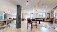 Located at 4573 Ponce de Leon Blvd in the vibrant city of Coral Gables, Florida, this single-floor communal living space offers a generously proportioned, open-plan layout designed for both social interaction and relaxed gathering. The interior is anchored by polished concrete floors that lend a contemporary, industrial-chic aesthetic throughout.

The space is thoughtfully divided into distinct yet connected zones. A social lounge area features an eye-catching green tile-clad structural column, a billiards table, and a dining nook set against a deep teal accent wall, illuminated by stylish pendant lighting. Warm wood paneling lines several walls, providing a natural contrast to the crisp white ceilings fitted with recessed lighting.

Separate seating areas are furnished with a curated mix of plush, rounded sofas and armchairs in rich terracotta, navy, and warm brown tones, arranged around low coffee tables atop vibrant area rugs. A gracefully curved wood partition wall adds architectural interest, subtly defining zones while maintaining the open flow of the space.

Large glass doors and windows draw in abundant natural light and provide direct access to an outdoor terrace, offering views of the surrounding Coral Gables streetscape. The overall ambiance is welcoming and design-forward, balancing modern sophistication with comfortable, communal warmth.