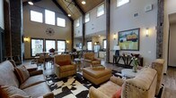 This remarkable single-floor space captivates from the moment you enter, boasting dramatic vaulted ceilings clad in warm wood paneling and anchored by bold, exposed structural beams that rise to impressive heights. Clerestory windows positioned high along the roofline flood the interior with abundant natural light, enhancing the open, airy atmosphere throughout. The open-concept layout connects the living, dining, and kitchen areas in a seamless flow, making the space feel expansive and highly functional. The living area is anchored by a cowhide area rug and furnished with rich leather armchairs and an upholstered ottoman, creating an inviting Western-inspired aesthetic. A large piece of artwork depicting a pastoral countryside scene adds a striking focal point near the entertainment console. The kitchen, visible from the main living space, features stainless steel appliances and classic cabinetry, while the adjacent dining area accommodates a generous table ideal for gathering. Industrial-style pendant lighting and wall sconces complement the rustic-modern design language throughout. Dark hardwood floors ground the space, providing a beautiful contrast to the warm tones of the leather furnishings and wooden architectural details. The overall ambiance is both sophisticated and welcoming, blending ranch-inspired charm with contemporary comfort.