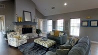 This single-floor home showcases a thoughtfully designed living room as its centerpiece. The space is immediately distinguished by its soaring vaulted ceilings, which lend an airy, open feel to the interior. The walls are finished in a cool, medium gray tone that establishes a modern and cohesive aesthetic throughout.

A striking stone fireplace serves as the room's focal point, featuring a natural stacked-stone surround and a dark mantel accented by a framed mirror above. The hearth adds both architectural character and warmth to the space.

Multiple windows along the walls flood the room with natural light, offering views to the exterior and enhancing the sense of openness. French doors near the fireplace provide an elegant transition to an adjacent area, adding to the room's connectivity and flow.

The living area is furnished with gray upholstered seating accented by floral and yellow throw pillows, complemented by a striped ottoman that doubles as a coffee table. Dark hardwood-style flooring contrasts with a plush charcoal area rug, grounding the seating arrangement and adding texture to the overall design.