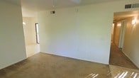 This single-floor space presents a straightforward, functional layout centered around an open living area. The main room features wall-to-wall beige carpeting, providing a warm and neutral foundation that pairs well with the clean white walls throughout. A ceiling-mounted light fixture serves as the primary overhead lighting source, while multiple electrical outlets are conveniently positioned along the walls.

An HVAC vent is integrated into the ceiling, ensuring climate control throughout the space. Natural light filters in from a sliding glass door or large window visible toward the rear of the room, which appears to open onto a balcony or outdoor area, adding a sense of connection to the exterior environment. The transition between the carpeted living area and what appears to be a tile or hard-surface flooring in the adjacent space is visible, suggesting distinct functional zones within the unit.

The overall ambiance is clean, minimal, and neutral, offering a blank canvas for personalization. The space is well-maintained and move-in ready, with simple finishes and an open feel suited to a variety of furniture arrangements and interior styles.