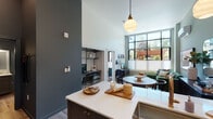 This thoughtfully designed single-floor residence at 4103 Mercantile Dr in Lake Oswego, Oregon, showcases a modern aesthetic anchored by soaring ceilings and an abundance of natural light. The open-concept living and kitchen area is a standout, featuring a bold slate-blue island with a quartz waterfall countertop, seating for multiple guests, and globe pendant lighting overhead. The kitchen is equipped with stainless steel appliances, a gas range, subway tile backsplash, and two-tone cabinetry in dark and light gray with brass hardware, complemented by a full-height refrigerator.

The living area is defined by dramatic floor-to-ceiling steel-framed windows that frame views of lush outdoor greenery, filling the space with sunlight. A dedicated nook off the main living area provides a functional workspace. The bedroom continues the window-forward design, offering generous natural light and access to a walk-in closet. The bathroom features a quartz vanity surface and slate-gray cabinetry. Throughout, light wood-look flooring, a mini-split HVAC system, and clean architectural lines contribute to a cohesive, contemporary atmosphere well-suited to modern living.