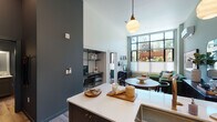 This thoughtfully designed single-floor residence at 4103 Mercantile Dr in Lake Oswego, Oregon, showcases a modern aesthetic anchored by soaring ceilings and an abundance of natural light. The open-concept living and kitchen area is a standout, featuring a bold slate-blue island with a quartz waterfall countertop, seating for multiple guests, and globe pendant lighting overhead. The kitchen is equipped with stainless steel appliances, a gas range, subway tile backsplash, and two-tone cabinetry in dark and light gray with brass hardware, complemented by a full-height refrigerator.

The living area is defined by dramatic floor-to-ceiling steel-framed windows that frame views of lush outdoor greenery, filling the space with sunlight. A dedicated nook off the main living area provides a functional workspace. The bedroom continues the window-forward design, offering generous natural light and access to a walk-in closet. The bathroom features a quartz vanity surface and slate-gray cabinetry. Throughout, light wood-look flooring, a mini-split HVAC system, and clean architectural lines contribute to a cohesive, contemporary atmosphere well-suited to modern living.