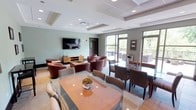 This single-floor amenity space is thoughtfully designed to serve as a versatile gathering and relaxation hub. The interior lounge area is finished with soft sage-green walls, recessed lighting, and a coffered ceiling that adds architectural depth to the room. A large wall-mounted flat-screen television anchors one end of the lounge, complemented by a warm wood media console and an arrangement of plush leather armchairs and a central coffee table — creating an inviting, residential atmosphere ideal for casual gatherings or quiet downtime.

The space transitions seamlessly into a dining area furnished with a glass-topped table and upholstered chairs, suitable for group meals or meetings. A dedicated kitchenette features granite countertops, rich dark wood cabinetry, a stainless steel sink, and full-height decorative tile backsplash, providing practical utility in an elegant setting.

Floor-to-ceiling windows and glass doors flood the interior with abundant natural light and open directly onto a generously sized outdoor terrace. The patio is outfitted with wicker lounge furniture featuring warm yellow cushions, surrounding low tables amid planters filled with seasonal flowers. Mature trees frame the terrace, providing a lush, green backdrop that enhances the sense of openness and connection to nature.