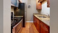 Located at 78 Manor in Manchester, New Hampshire, this single-floor unit offers a clean, functional layout with consistent warm-toned hardwood flooring throughout. The open living area welcomes you upon entry, bathed in natural light from a large window that frames views of lush greenery outside. The space flows naturally toward the galley-style kitchen, which is well-appointed with rich cherry wood cabinetry, granite countertops, a stainless steel refrigerator, electric range, and built-in microwave — all complemented by a wall-mounted ventilation fan. The bedrooms are generously sized, each featuring the same gleaming hardwood floors, neutral beige walls, and individual windows that bring in ample natural light and outdoor views. Baseboard heating runs along the perimeter of the rooms, with a wall-mounted air conditioning unit providing year-round climate control. The bathroom features classic white subway-style wall tiling surrounding a full soaking tub, offering a clean and timeless aesthetic. Throughout the unit, crisp white baseboards provide a polished contrast to the warm flooring and neutral walls, creating a bright, cohesive ambiance that is both comfortable and move-in ready.