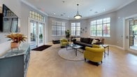 This single-floor community space offers a thoughtfully designed interior that balances functionality with contemporary style. The main lounge area serves as the heart of the space, featuring large windows with horizontal blinds that invite abundant natural light, elegant crown molding, and large-format tile flooring. A set of glass-paneled double French doors with a transom window above opens to the exterior, creating a seamless indoor-outdoor connection. The lounge is furnished with a comfortable sectional sofa, distinctive chartreuse accent chairs, a round area rug, and glass coffee tables, all anchored beneath a pendant light fixture. Recessed lighting and neutral gray walls contribute to the open, airy atmosphere throughout.

An entry area adjacent to the lounge provides additional seating, wall-mounted media, and a decorative console, offering a warm welcome upon arrival. The fully equipped kitchen features rich dark cabinetry paired with granite countertops showcasing bold, multicolored veining. A gray subway tile backsplash, stainless steel appliances including a refrigerator, dishwasher, and over-the-range microwave, and a double basin sink complete this practical and attractive culinary space. Frosted glass upper cabinet doors add a refined touch. The overall ambiance of the space is polished, inviting, and well-suited for gatherings and community use.
