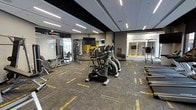 This contemporary fitness center offers a spacious, single-floor layout designed to accommodate a full range of workout activities. The space features a sleek, modern aesthetic with a dark suspended ceiling accented by rows of linear LED lighting that illuminate the floor evenly. Patterned carpet tiles in grey and gold tones cover the floor, adding a stylish touch to the functional environment.

The cardio zone is prominently featured at the center of the space, housing multiple elliptical trainers and treadmills arranged for easy access and ample spacing between machines. A large flat-screen television is mounted on the wall, providing entertainment during workouts. The strength training area is equipped with a variety of cable and resistance machines, along with free weight stations, catering to users of all fitness levels.

Glass partitions and walls on multiple sides create a sense of openness while defining the space, allowing natural light to filter through and offering views into adjacent areas. Bold yellow accent walls add a vibrant, energetic contrast to the otherwise neutral color palette. The overall ambiance is clean, professional, and motivating, reflecting a high-quality facility suited for both casual exercisers and dedicated athletes.