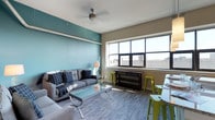 This single-floor space makes a strong visual impression through its thoughtful use of color and light. The living area is anchored by a bold teal accent wall that contrasts beautifully against the neutral taupe tones of the surrounding walls, creating a dynamic and contemporary atmosphere. Large, multi-pane windows flood the room with natural light and offer open views of the surrounding urban skyline, enhancing the sense of space and connectivity to the city outside. The wood-style flooring in a cool grey finish runs throughout, adding warmth and cohesion to the overall aesthetic. A ceiling fan with an industrial-inspired fixture provides both function and a subtle design detail overhead. The living area comfortably accommodates a full sofa set paired with a sleek glass coffee table, while a console table positioned beneath the windows serves as both a functional surface and a visual anchor to the window wall. Bar-height stools in a vivid lime green introduce a playful pop of color, complementing the teal tones and reinforcing the space's modern, energetic character. Overall, the space strikes a balance between urban edge and comfortable livability, making it both visually engaging and highly functional.