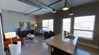 This single-floor urban loft showcases a compelling blend of industrial and contemporary design. The open-concept layout seamlessly connects the kitchen, dining, and living areas into one cohesive, flowing space. Overhead, exposed metallic ductwork and pendant lighting with an industrial flair add striking visual character to the high ceilings, reinforcing the loft's urban aesthetic.

The kitchen features a generous granite countertop island with an undermount sink, providing ample prep space and a natural gathering point. Dark lower cabinetry grounds the kitchen and complements the neutral gray tones found throughout the space. Pendant lights hang overhead, offering both task lighting and a stylish focal point.

Polished concrete floors extend throughout the entire space, contributing to the loft's modern, industrial character while remaining easy to maintain. The living area is well-lit thanks to multiple large windows that invite abundant natural light and offer views of the surrounding neighborhood. A colorful piece of wall art adds a vibrant contrast to the otherwise cool, neutral palette.

The overall ambiance is contemporary and urban — open, airy, and thoughtfully designed to balance functional living with aesthetic appeal, making it an ideal space for both relaxing and entertaining.