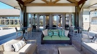 This single-level outdoor pavilion serves as a sophisticated communal gathering space, seamlessly blending covered comfort with open-air living. The structure is defined by its striking timber-frame construction, featuring exposed wood beams and a vaulted ceiling that lend a warm, craftsman-inspired aesthetic. At the heart of the space stands a floor-to-ceiling stacked stone fireplace, complemented by a wall-mounted flat-screen television, creating an inviting focal point ideal for relaxation and entertainment. The pavilion is finished with durable slate tile flooring throughout, providing a cohesive and elegant foundation. Generously arranged lounge seating, including wicker sofas and upholstered armchairs with coordinating accent pillows, encourages comfortable gathering in multiple zones. Expansive arched openings on multiple sides allow abundant natural light to flood the space while framing unobstructed views of the resort-style swimming pool and the surrounding courtyard and residential buildings. A set of full-length French doors connects the pavilion to an adjacent interior amenity space, further enhancing the indoor-outdoor flow. Dining tables and bar-height seating extend the usable area into the open patio beyond, accommodating a variety of social settings throughout the day.