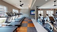 Located at 3100 Merritt Rd in Rowlett, TX, this single-floor fitness center is a thoughtfully designed facility offering a comprehensive workout environment. The space is organized into distinct functional zones, each dedicated to a different type of exercise. The cardio area features a long row of treadmills positioned alongside large windows, providing views of the surrounding grounds and filling the space with natural light. Stationary bikes and elliptical machines complement the cardio offerings. A dedicated strength training section houses a variety of cable machines, multi-station weight equipment, and free weight benches, supported by floor-to-ceiling mirrors that enhance the sense of openness. A separate open-floor stretching and mat area, painted in a distinctive blue accent, provides ample room for flexibility work or bodyweight exercises. Throughout the facility, the flooring showcases a striking multi-tone pattern combining light, dark, and warm-toned tiles, adding visual interest to the space. Ceiling fans and recessed lighting maintain a comfortable atmosphere, while multiple flat-screen TVs are mounted throughout. Large glass doors and expansive windows ensure connectivity to the outdoors, making this fitness center a bright, energizing, and fully equipped space for a wide range of workout routines.