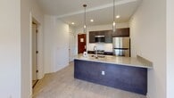 Located on the historic Monument Avenue in Richmond, Virginia, this single-floor residence combines modern design with functional living spaces. The open-concept kitchen serves as a central highlight, featuring dark espresso cabinetry, gleaming granite countertops, a bold navy-accented island with an undermount sink, and a full suite of stainless steel appliances including a refrigerator, range, and microwave. White subway tile backsplash adds a crisp, classic touch, while pendant lighting overhead creates a warm, inviting atmosphere. Light wood-style flooring flows through the main living areas, contributing to a clean and contemporary aesthetic. The bedroom offers a quiet retreat with plush carpeting, crisp white walls, and architectural ceiling details that add visual interest to the space. The bathroom is finished with neutral subway tile in the walk-in shower, chrome fixtures, and a clean, minimalist design. Recessed lighting throughout provides consistent illumination, and the overall layout is thoughtfully arranged to maximize both comfort and functionality. The residence reflects a refined, modern sensibility well-suited to its iconic Richmond address.