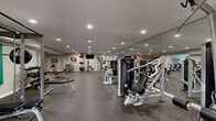 Located at 1900 Tree Mountain Parkway in Stone Mountain, Georgia, this well-appointed fitness center offers a generously sized, single-level layout designed to accommodate a full spectrum of workout needs. The space is finished with durable rubber flooring throughout, providing both safety and comfort underfoot, while recessed lighting illuminates the entire area evenly and effectively.

The strength training section is extensively equipped with multiple selectorized cable machines, adjustable benches, and a full rack of color-coded kettlebells. A tiered set of stability and medicine balls adds functional training versatility, and floor-to-ceiling mirrors along one wall enhance the sense of space while allowing users to monitor their form.

The cardio zone features treadmills, ellipticals, recumbent bikes, and indoor cycling bikes, with motivational fitness artwork displayed along the walls. A wall-mounted flat-screen television serves the strength area for added convenience.

Natural light fills a dedicated section of the facility through large windows and a glass door that opens directly to an outdoor pool area, creating an energizing connection to the lush, tree-lined surroundings. The neutral gray walls and clean, modern aesthetic give the entire fitness center a professional yet welcoming ambiance suited for residents of all fitness levels.
