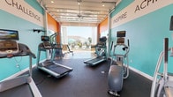 Located at 290 Bagwell Rd in Greenville, SC, this single-level fitness center is designed to energize and motivate with its bold, well-considered aesthetic. The interior is defined by striking teal and orange walls adorned with large motivational words—"Inspire," "Challenge," "Commit," "Play," "Define," "Stretch," and "Motivate"—creating a dynamic and encouraging atmosphere throughout the space.

The main workout area is equipped with multiple treadmills, elliptical trainers, a rowing machine, stationary bikes, and strength equipment including a multi-use cable station and pull-up bar. Rubber flooring throughout provides safety and durability underfoot. Ceiling fans ensure consistent air circulation during workouts.

A standout architectural feature is the full-width glass garage door on one end of the space, which opens completely to reveal a direct, unobstructed view of an outdoor pool and patio area. This integration of indoor and outdoor environments allows natural light to flood the room and creates a seamless connection to the surrounding amenity space.

On the opposite end, a set of French doors flanked by large windows further enhances natural light and provides additional views of the outdoor surroundings. Recessed lighting supplements natural illumination throughout the day, ensuring the space remains bright and functional at all hours.