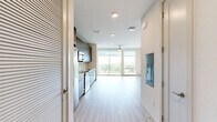 This single-story unit offers a thoughtfully designed open-concept layout that maximizes its compact footprint. Light wood-style flooring runs throughout, creating a cohesive and airy feel that connects each area seamlessly.

The combined kitchen and living area serves as the heart of the unit. The kitchen is outfitted with floor-to-ceiling dark wood cabinetry providing ample storage, white quartz countertops, a white tile backsplash, and a full suite of stainless steel appliances including a refrigerator, electric range, built-in microwave, and dishwasher. The living area benefits from large floor-to-ceiling windows and a sliding glass door that opens to a balcony, flooding the space with natural light and offering open outdoor views of surrounding greenery and neighboring buildings. A ceiling fan adds practical comfort to the living space.

The hallway connects the main living area to the bathroom and includes a closet with a hanging rod for storage. The bathroom features neutral gray tile flooring, a warm wood-toned vanity with a white countertop, and clean, textured white walls, maintaining the unit's modern, understated aesthetic throughout.