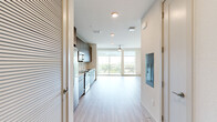 This single-story unit offers a thoughtfully designed open-concept layout that maximizes its compact footprint. Light wood-style flooring runs throughout, creating a cohesive and airy feel that connects each area seamlessly.

The combined kitchen and living area serves as the heart of the unit. The kitchen is outfitted with floor-to-ceiling dark wood cabinetry providing ample storage, white quartz countertops, a white tile backsplash, and a full suite of stainless steel appliances including a refrigerator, electric range, built-in microwave, and dishwasher. The living area benefits from large floor-to-ceiling windows and a sliding glass door that opens to a balcony, flooding the space with natural light and offering open outdoor views of surrounding greenery and neighboring buildings. A ceiling fan adds practical comfort to the living space.

The hallway connects the main living area to the bathroom and includes a closet with a hanging rod for storage. The bathroom features neutral gray tile flooring, a warm wood-toned vanity with a white countertop, and clean, textured white walls, maintaining the unit's modern, understated aesthetic throughout.