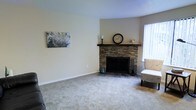 This single-floor home offers a welcoming and comfortable living environment defined by clean lines and warm, neutral tones. The living area serves as the heart of the space, centered around a standout stacked-stone fireplace with a rich, dark wood mantel — a focal point that adds both texture and warmth to the room. The hearth is finished with slate-toned tile, providing a durable and stylish contrast to the plush, light-colored carpeting that extends throughout the space. The walls are painted in a soft, creamy white that enhances the sense of openness and reflects natural light effectively. A window positioned adjacent to the fireplace allows natural light to filter into the room, contributing to its bright and airy feel. The layout is open and spacious, offering flexibility for a variety of furniture arrangements. The overall ambiance of this home is calm and inviting, with architectural details — particularly the stone fireplace — lending a sense of character and craftsmanship. The single-floor layout ensures easy accessibility and a seamless flow throughout the home, making it a practical and appealing living space.