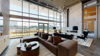 Located at 417 W Peace St in Raleigh, NC, this sophisticated lounge space is designed to impress with its contemporary architecture and high-end finishes throughout. The single-floor layout is anchored by dramatic double-height ceilings and an expansive floor-to-ceiling curtain wall of glass that floods the interior with natural light and offers sweeping views of the surrounding cityscape. A standout feature is the striking full-height marble fireplace surround, which serves as the room's focal centerpiece, flanked by curated seating areas with plush leather sofas and modern accent chairs. An adjacent media lounge showcases a boldly designed diagonal wood-slat accent wall framing a large flat-screen television, paired with large-format tile flooring and additional lounge seating. The space transitions seamlessly to an outdoor terrace through a set of glass double doors, where alfresco seating beneath umbrellas extends the living area into the open air. Interior details include a slatted wood ceiling treatment, globe cluster pendant lighting, and contemporary artwork on display. A natural wood sliding barn door adds a warm, organic contrast to the otherwise sleek modern palette. The overall ambiance is refined yet inviting, balancing luxury materials with comfortable, functional design.