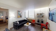 This single-floor residence offers a thoughtfully arranged layout across several distinct living spaces. The living room serves as the heart of the home, featuring rich hardwood-style flooring, a sleek black leather sofa, and vibrant red accent chairs. An open shelving unit displays curated décor, while a large window with blinds fills the room with natural light. A flat-screen TV and vintage-style posters, including a classic Monaco Grand Prix print, add personality to the space.

The compact kitchen is efficiently designed with white cabinetry, dark countertops, classic white subway tile backsplash, and essential appliances including an electric range and sink — a practical and clean workspace.

Two well-sized bedrooms provide comfortable retreats. The primary bedroom features a striking four-poster bed frame, warm bedside lighting, and a mirrored wardrobe, creating an elegant atmosphere. The second bedroom offers carpeted flooring, a study desk with natural light from a large window, and a relaxed, functional layout suited for work or rest.

The bathroom features a standout yellow vanity countertop paired with a white basin sink, a wall-mounted toilet in an adjacent water closet, built-in shelving, and a mirrored wall — combining practicality with a pop of color throughout.