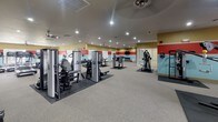 This spacious, single-floor fitness center is thoughtfully designed to accommodate a full range of workout activities across multiple interconnected zones. The interior is immediately recognizable by its distinctive tri-color wall scheme — bands of soft yellow, bold red, and light blue — which lend the space a vibrant, energetic atmosphere throughout.

The cardio area features a generous row of treadmills and elliptical machines aligned along wide windows that offer outdoor views, filling the room with natural light. Ceiling fans are positioned throughout to maintain airflow and comfort during workouts.

The strength training section is well-stocked with a variety of Cybex cable machines and multi-station weight equipment, arranged with ample space between units for safe use. Full-length mirrors along one wall visually expand the area and allow users to monitor their form.

A separate, glass-partitioned room serves as a dedicated group fitness or activity space. It features a wall-mounted television, a decorative console, and exercise balls, making it suitable for stretching, yoga, or guided classes.

Recessed lighting throughout ensures consistent illumination across all areas, while the neutral carpeted flooring and rubber equipment mats contribute to a clean, functional aesthetic. The overall layout offers a comprehensive and accessible fitness experience.