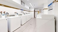Located at 160 Erskine Ave in Toronto's Midtown neighbourhood, this shared laundry facility offers a clean, functional, and well-organized environment for residents. The space is divided into two interconnected areas, together providing a generous footprint for laundry operations.

The main laundry room features two long rows of commercial-grade top-loading washing machines and front-loading dryers, all finished in white, and numbered for easy identification. The machines are equipped with digital payment terminals, offering a convenient card-based payment system. Bright fluorescent linear lighting runs across the ceiling, ensuring excellent visibility throughout.

The walls are dressed in white tile adorned with a decorative gold horizontal border, lending a clean and polished aesthetic. Light grey large-format floor tiles complement the overall neutral palette. A wall-mounted mini-split air conditioning unit ensures year-round comfort.

An adjoining area serves as a utility and prep zone, featuring a granite-topped folding counter with open shelving below — stocked with a curated collection of books, adding a thoughtful community touch. A stainless steel utility sink provides added functionality. A payment kiosk station is also present for added convenience. Small hopper-style windows allow for passive ventilation, while a security camera ensures the safety of the space.