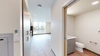Located at 1411 E Iron Brg Way in Spokane, WA, this efficiently designed unit offers a well-organized single-floor layout comprising a living area, kitchen, bathroom, and hallway. The interior features light wood-tone vinyl flooring throughout, crisp white walls, and recessed lighting that contribute to a clean, modern aesthetic.

The open living area is anchored by a large floor-to-ceiling window that invites abundant natural light and provides views of the surrounding cityscape. A wall-mounted mini-split heating and cooling unit ensures year-round climate comfort. Built-in gray cabinetry along the hallway offers convenient storage solutions.

The kitchen is a standout feature, outfitted with two-tone cabinetry — lower gray cabinets paired with bold blue upper cabinets — and stainless steel appliances including a range, microwave, refrigerator, and dishwasher. Light countertops and an additional large window further brighten the space.

The bathroom is thoughtfully appointed with gray cabinetry and a vanity, and notably includes a stacked washer and dryer unit, adding full in-unit laundry convenience. The hallway connects all areas efficiently, with warm wood-framed doorways adding a subtle design accent throughout the unit.