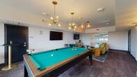 Located at 5745 Golden Triangle Blvd in Fort Worth, Texas, this single-floor amenity lounge is a well-appointed recreational space designed for social gatherings and leisure. The layout is open and generously proportioned, anchored by a striking pool table with a vibrant teal felt surface and a solid wood frame, positioned beneath two eye-catching starburst chandeliers with brass finishes that lend a mid-century modern aesthetic to the room. Large-format wall-mounted televisions are installed throughout, enhancing the entertainment experience. A shuffleboard table is positioned near a wall of oversized windows that frame views of the surrounding Fort Worth landscape, filling the space with abundant natural light. Textured accent walls add depth and visual interest to the room's design. The lounge area features plush seating including a velvet gold sofa and teal accent chairs, complemented by glass-top coffee tables with gold frames and layered area rugs in rich, jewel-toned hues. Decorative floral artwork in dark-framed panels adds a dramatic focal point alongside bistro-height tables for casual seating. A light wood media credenza with circular brass hardware grounds the TV lounge area. The overall ambiance is vibrant, modern, and inviting, balancing bold design choices with comfortable, functional furnishings.
