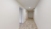 This single-floor unit presents a bright, minimalist design characterized by crisp white walls and light wood-look flooring that runs consistently throughout the space, creating a cohesive and airy atmosphere. The layout, visible in the overhead floor plan, is efficiently organized with clearly defined areas connected by a central corridor.

The main living and entry area is illuminated by recessed lighting and equipped with a wall-mounted mini-split air conditioning unit, ensuring climate control year-round. A set of double closet doors along the hallway provides convenient built-in storage. The entry leads to a well-lit front door area that continues the clean, uncluttered aesthetic.

The kitchen is outfitted with gray cabinetry, white subway tile backsplash, white appliances including an electric range and built-in microwave, and a single-basin sink — offering a functional and tidy cooking space.

The bedroom receives natural light through a window fitted with blinds, offering a quiet and comfortable retreat with ample open floor space.

The bathroom features wood-look tile flooring, a gray vanity with under-sink storage, a wall-mounted light fixture above the mirror, a toilet, and a step-in shower with tiled walls, maintaining the unit's clean, consistent palette throughout.