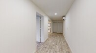 This single-floor unit presents a bright, minimalist design characterized by crisp white walls and light wood-look flooring that runs consistently throughout the space, creating a cohesive and airy atmosphere. The layout, visible in the overhead floor plan, is efficiently organized with clearly defined areas connected by a central corridor.

The main living and entry area is illuminated by recessed lighting and equipped with a wall-mounted mini-split air conditioning unit, ensuring climate control year-round. A set of double closet doors along the hallway provides convenient built-in storage. The entry leads to a well-lit front door area that continues the clean, uncluttered aesthetic.

The kitchen is outfitted with gray cabinetry, white subway tile backsplash, white appliances including an electric range and built-in microwave, and a single-basin sink — offering a functional and tidy cooking space.

The bedroom receives natural light through a window fitted with blinds, offering a quiet and comfortable retreat with ample open floor space.

The bathroom features wood-look tile flooring, a gray vanity with under-sink storage, a wall-mounted light fixture above the mirror, a toilet, and a step-in shower with tiled walls, maintaining the unit's clean, consistent palette throughout.