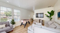 Located in Vista, CA, this single-floor club room offers a well-appointed, open-concept layout that seamlessly combines a kitchen and living area into one inviting, communal space. The interior is finished with light-toned hardwood flooring and crisp white walls, creating a bright, airy atmosphere throughout.

The kitchen is outfitted with full-height white shaker cabinets, sleek marble-look quartz countertops, stainless steel appliances including a refrigerator and microwave, and a deep single-basin sink — offering a fully functional setup for entertaining. Recessed lighting ensures the space is well-lit at all times.

The living area is generously furnished with multiple seating arrangements, including a large plush sectional sofa and an additional tufted gray sectional, providing ample comfort for gatherings. A wall-mounted flat-screen TV anchors the main lounge area, complemented by a natural wood media console and a tall tropical plant that adds a refreshing organic touch. Ocean-inspired artwork adorns the walls, reinforcing a relaxed coastal aesthetic.

A foosball table adds a recreational dimension to the space, making it ideal for social events. Large windows along one wall invite abundant natural light and outdoor views, while a glass door opens directly to a pool area, enhancing the club room's resort-like appeal. A bistro-style high-top table near the entrance offers an additional casual seating option.