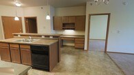 This single-story space showcases an open-concept kitchen that connects naturally to the surrounding living areas. The kitchen is outfitted with medium-toned wood cabinetry that lines the walls, offering generous storage through both upper and lower cabinet configurations. A built-in wine rack integrated into the upper cabinetry adds a functional decorative touch. The countertops feature a neutral laminate finish that complements the warm wood tones throughout.

A peninsula with a sink and black dishwasher defines the kitchen boundary, providing both workspace and a subtle division between the kitchen and the adjoining areas. A pendant light fixture hangs above, supplemented by a chandelier visible in the adjacent room, contributing to a layered lighting scheme.

A dedicated built-in desk area with drawers sits along the far wall, creating a convenient workspace within the kitchen zone. The kitchen floor features durable tile in a stone-like neutral pattern, which transitions to carpet in the neighboring rooms. Doorways on either side of the kitchen provide access to additional spaces, reinforcing the open and connected layout of this single-floor residence.